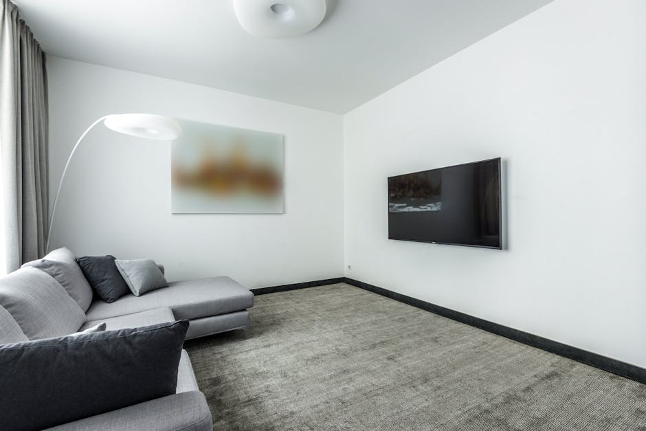 A modern living room featuring a light grey sofa with multiple cushions, situated on a textured grey carpeted floor. To the left, a tall, curved white floor lamp with a circular lampshade illuminates 
